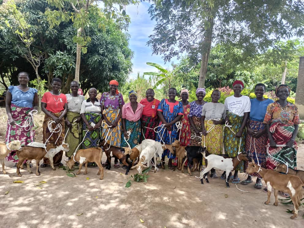 A group of women standing together outdoors, each holding a goat on a leash, with lush greenery in the background.