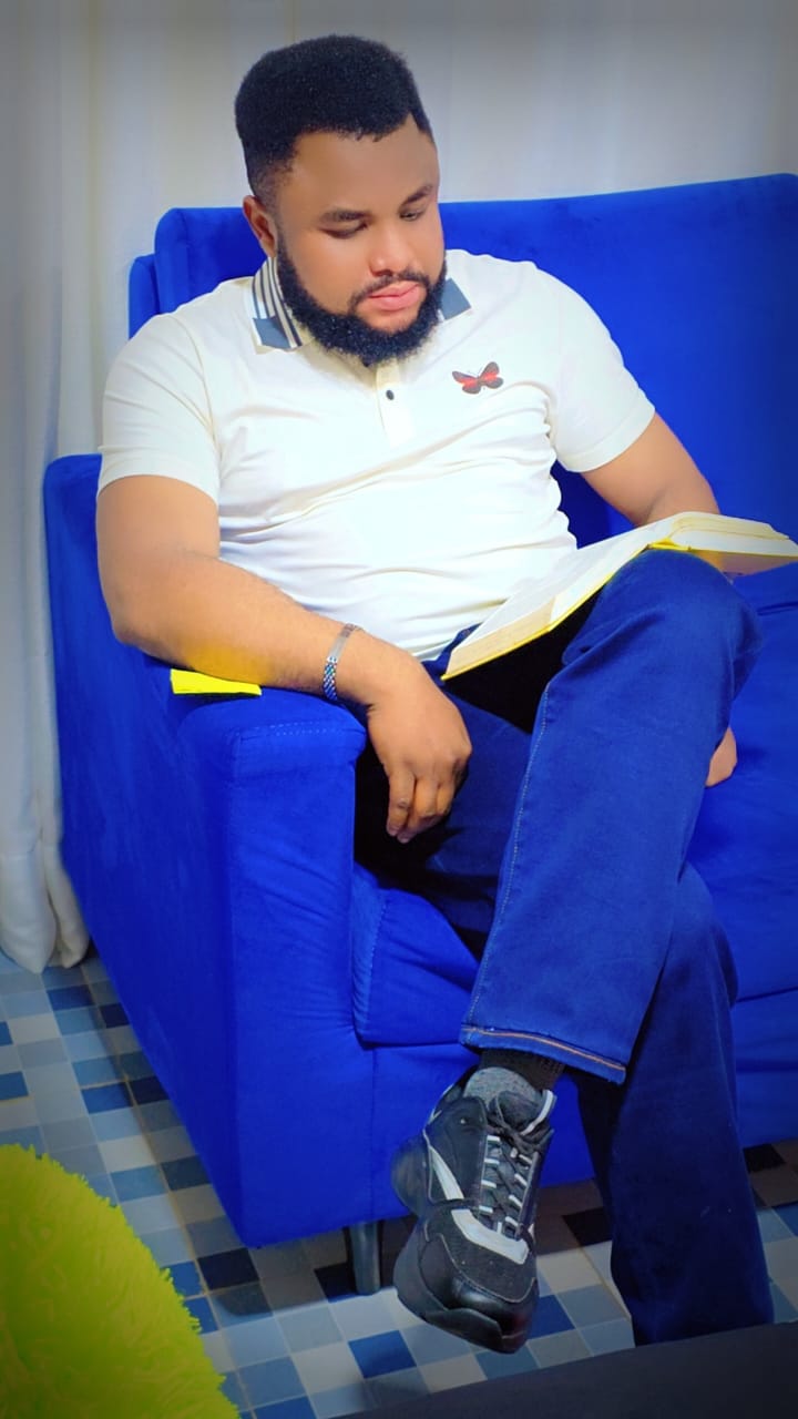 A person reading a book while sitting on a blue couch, dressed in a white polo shirt with a butterfly emblem and dark blue pants.